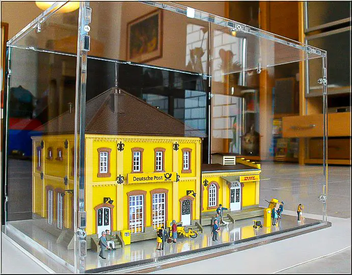 Acrylic display case over a model railway station with a yellow Deutsche Post facade and small figures on the platform